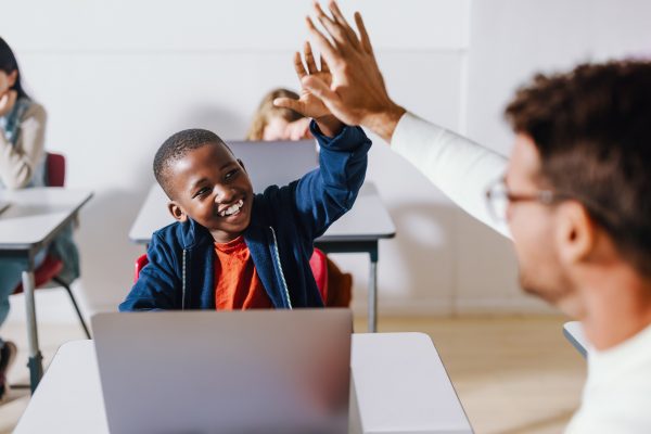 Happy kid high fiving his teacher in a computer science class, celebrating successful teaching and learning. Student getting attention and assistance from his teacher as he learns digital literacy.; Shutterstock ID 2265470543; purchase_order: 978-0-19-457154-8; job: Assistant Marketing Manager; client: ELT; other: